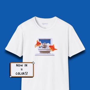 May include: A white t-shirt featuring a graphic of a vintage computer monitor displaying a mouse against a blue sky with clouds, flanked by two orange goldfish. A rectangular sign in the corner displays the text NOW IN 11 COLORS!