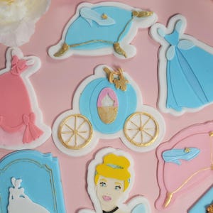 May include: A collection of Cinderella-themed decorations, including a carriage, dresses, and Cinderella's likeness. The decorations are in shades of blue, pink, and gold, with white accents, set against a pink background.