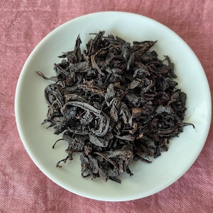 May include: A pile of dark brown, dried, twisted tea leaves centered on a small, round, white ceramic plate. The leaves have a matte, textured appearance with varying shades of deep brown and black, showing curled and folded edges throughout.