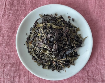 May include: A pile of loose-leaf tea consisting of a blend of dark brown, nearly black, and muted green dried leaves. The tea is contained within a simple, smooth, white ceramic saucer, presenting a mix of curled and fragmented botanical pieces.