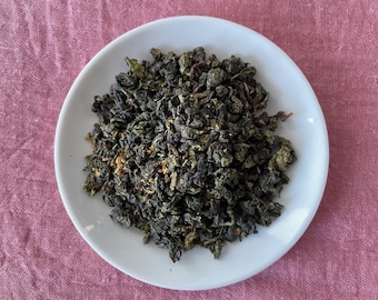 May include: A pile of dark green, tightly rolled oolong tea leaves with small flecks of yellow and brown botanical matter, centered on a plain white circular ceramic saucer. The tea leaves have a textured, crinkled appearance and are dry.