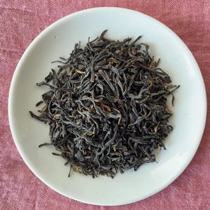 May include: A pile of loose-leaf black tea sits in the center of a small, round, white ceramic saucer. The tea leaves are dark brown to black in color, with some lighter golden-brown tips visible throughout the mixture of curled and twisted foliage.