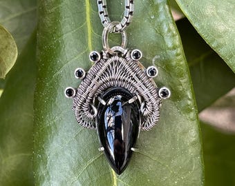 May include: A silver chain necklace with a black gemstone pendant. The pendant is surrounded by a silver wire design and small black gemstones.
