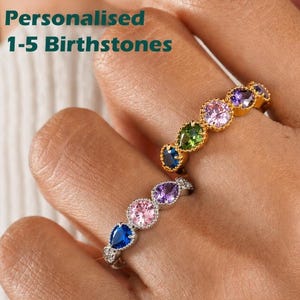 May include: Two rings featuring multiple round and pear-shaped gemstones in various colors including blue, pink, green, and purple. The text 'Personalised 1-5 Birthstones' is visible. The rings have metallic bands with a beaded or textured edge.