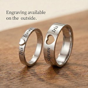 Op de afbeelding: Twee zilveren ringen met gegraveerde namen en hartdetails. Eén ring heeft een hartvormige uitsparing, de andere heeft een klein hartje. De tekst "Graveren aan de buitenkant mogelijk" is zichtbaar.
