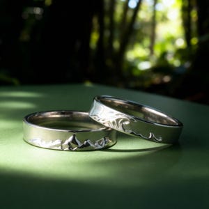 May include: Two silver rings with nature-inspired designs. One ring features a mountain range, and the other depicts ocean waves. The rings are set against a green background, with sunlight creating shadows.