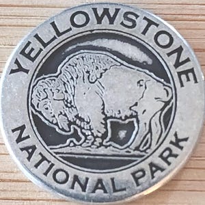 May include: A silver-colored Yellowstone National Park souvenir coin. The coin features a detailed image of a bison in the center, surrounded by the words "YELLOWSTONE NATIONAL PARK" in a circular pattern.