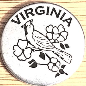 May include: A round, silver-colored souvenir featuring the word "VIRGINIA" in a curved font. A black and white cardinal bird is perched on a flowering branch. The design is a state emblem.
