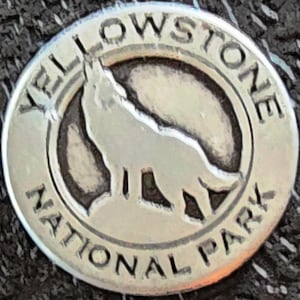 May include: A silver-colored round Yellowstone National Park souvenir pin. The pin features a howling wolf silhouette inside a dark circle, with the words "YELLOWSTONE NATIONAL PARK" around the edge.