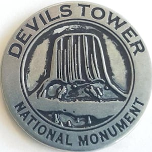 May include: A silver-colored metal coin with a black and white image of Devils Tower National Monument. The coin has the words "Devils Tower National Monument" around the edge.