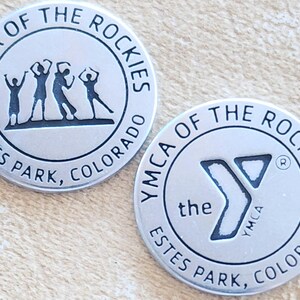 May include: Two silver metal tokens with the YMCA of the Rockies logo. The first token features a group of four people with their arms raised in the air. The second token features the YMCA logo with the text "the YMCA" and "Estes Park, Colorado".