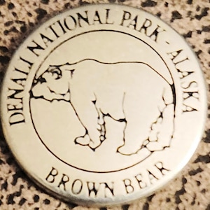 May include: A silver round button with the text "Denali National Park - Alaska" and "Brown Bear" around the edge. A brown bear is depicted in the center.