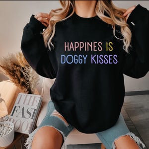 Op de afbeelding: Zwart sweatshirt met de tekst "Happiness is Doggy Kisses" in regenboogkleuren.