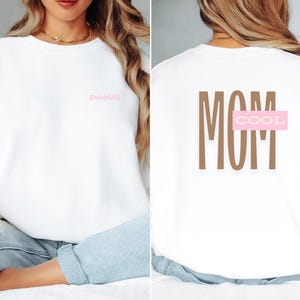 Peut inclure: Sweat-shirt blanc avec le mot "MOM" en lettres brunes et le mot "COOL" dans un rectangle rose au dos. Le devant du sweat-shirt porte le mot "EMMANUEL" en lettres roses.