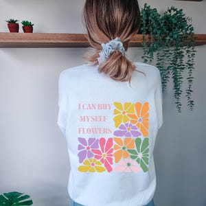 May include: White sweatshirt featuring the text "I CAN BUY MYSELF FLOWERS" and a colourful floral design. The design includes pink, orange, yellow, and green flowers. A comfortable, casual top.