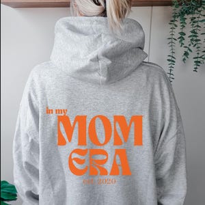 May include: Grey hooded sweatshirt with the text "in my MOM ERA est. 2020" printed in orange on the back.