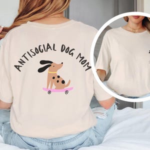 Peut inclure: Un t-shirt beige avec le texte "Antisocial Dog Mom" et un chien de dessin animé faisant du skateboard au dos.