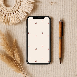Könnte beinhalten: Ein schwarzes Smartphone mit weißem Hintergrund und einem Muster aus roten Kirschen. Das Telefon liegt auf einer beigefarbenen Oberfläche mit einem braunen Stift daneben.