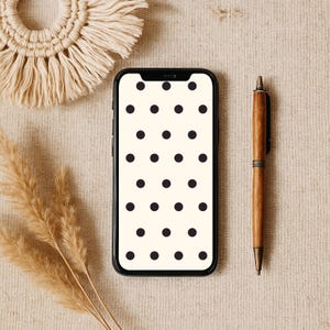 Può includere: Uno smartphone nero con uno sfondo bianco e puntini neri visualizzati sullo schermo. Il telefono è appoggiato su una superficie beige con una penna marrone e un oggetto intrecciato beige.