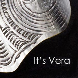 Może przedstawiać: Zbliżenie srebrnego, misternie rzeźbionego metalowego przedmiotu o teksturowanej powierzchni. Projekt zawiera zakrzywione linie i wzory. Tekst "It's Vera" jest wyświetlany białą czcionką na czarnym tle.