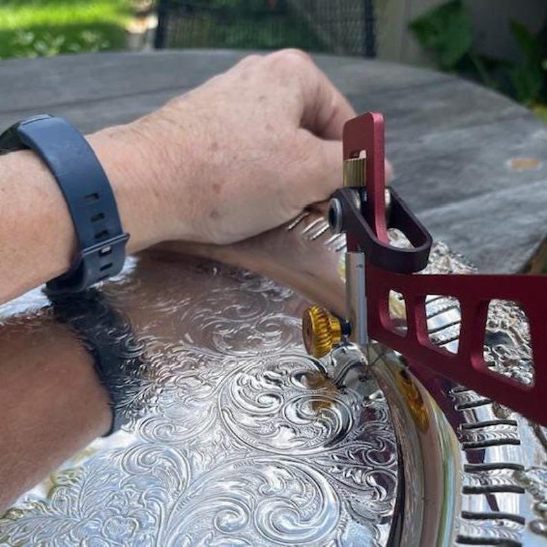 Może przedstawiać: Srebrny p&oacute;łmisek z ozdobnym kwiatowym wzorem. Czerwono-czarne metalowe narzędzie jest umieszczone na tacy. Ciemnoniebieski zegarek jest na nadgarstku.