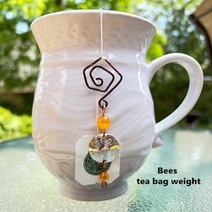 May include: A white ceramic mug with a tea bag weight attached to the string. The weight features a dark copper wire spiral, a yellow bead, and a silver-toned circular charm embossed with bee imagery, finished with another small yellow bead.