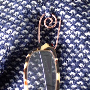 Peut inclure: Un porte-lunettes en fil de cuivre avec un motif en spirale, attaché à une paire de lunettes de lecture à motif écaille de tortue. Les lunettes sont posées sur un tissu en tricot bleu et blanc texturé.