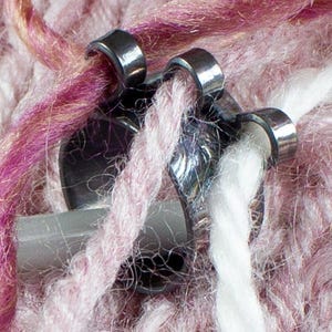 May include: Close-up of a metal zipper with a grey slider, holding together strands of pink, purple, and white yarn. The yarn has a textured, fuzzy appearance, suggesting a knitted or crocheted item. The zipper's metal components are shiny.