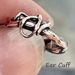 May include: An ear cuff featuring a textured, dark oxidized metal base with polished, copper-toned highlights. The design consists of an open, irregular loop structure with a rugged, organic appearance. The text Ear Cuff is visible.