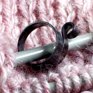 Może przedstawiać: Zbliżenie srebrnego znacznika rzęd&oacute;w na drutach do rob&oacute;tek ręcznych, na tle r&oacute;żowej dzianiny. Znacznik rzęd&oacute;w ma dekoracyjny, wirujący wz&oacute;r.