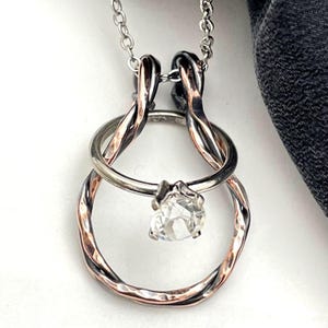 May include: A silver necklace with a pendant featuring a clear, faceted gemstone. The pendant has a unique design with intertwined metal rings in silver and copper tones, suspended from a delicate chain.