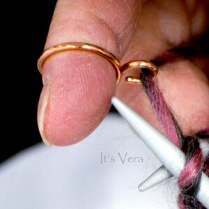 Może przedstawiać: Zbliżenie miedzianego pierścienia prowadzącego przędzę na palcu, z dołączonym drugim pierścieniem. Pierścień służy do robienia na drutach i szydełkowania. Na zdjęciu widać r&oacute;wnież druty do robienia na drutach i przędzę w odcieniach r&oacute;żu, czerni i bieli. Widoczny jest tekst "It's Vera".