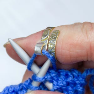 May include: Close-up of knitting needles with blue yarn and three silver and gold-toned knitting rings on a finger. The rings have decorative patterns. The yarn is being knitted with the needles.