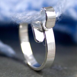 Może przedstawiać: Zbliżenie srebrnego pierścionka o unikalnym wzorze, kt&oacute;ry wydaje się trzymać pasmo jasnoniebieskiej przędzy. Pierścionek ma zakrzywiony, otwarty design z polerowanym wykończeniem, na rozmytym niebieskim tle.