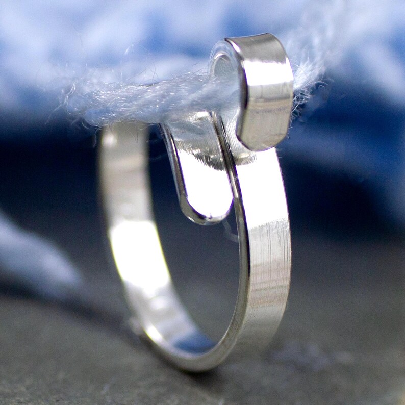 Może przedstawiać: Zbliżenie srebrnego pierścionka o unikalnym wzorze, kt&oacute;ry wydaje się trzymać pasmo jasnoniebieskiej przędzy. Pierścionek ma zakrzywiony, otwarty design z polerowanym wykończeniem, na rozmytym niebieskim tle.