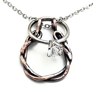 May include: A silver and copper pendant necklace with a clear, faceted gemstone. The pendant has a unique design with a ring and intertwined metalwork, suspended from a delicate chain. The gemstone is set in a silver prong setting.