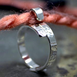 Może przedstawiać: Zbliżenie srebrnego pierścionka z teksturowanym paskiem i zakrzywionym wzorem. Pierścionek jest owinięty wok&oacute;ł pasma r&oacute;żowej przędzy, z małym srebrnym zapięciem. Pierścionek jest bardzo wypolerowany i odbija światło.