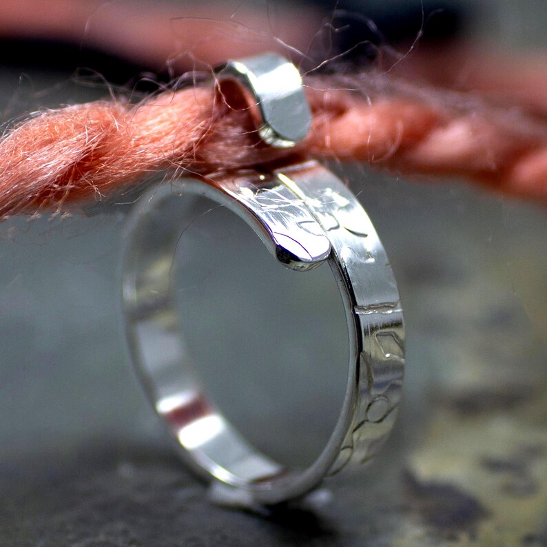 Może przedstawiać: Zbliżenie srebrnego pierścionka z teksturowanym paskiem i zakrzywionym wzorem. Pierścionek jest owinięty wok&oacute;ł pasma r&oacute;żowej przędzy, z małym srebrnym zapięciem. Pierścionek jest bardzo wypolerowany i odbija światło.