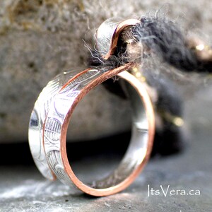 Może przedstawiać: Ręcznie robiony pierścionek ze srebrną obrączką i miedzianymi krawędziami. Srebrna obrączka ma skomplikowane grawerowane wzory. Pierścionek jest pokazany z bliska, podkreślając szczeg&oacute;łowe rzemiosło i kontrast między metalami.