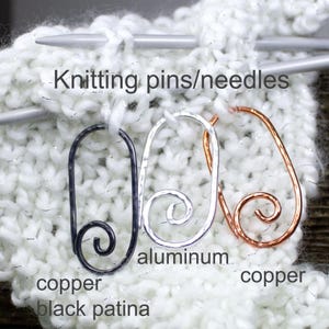 May include: Close-up of three knitting pins/needles. One is black copper with a black patina, one is aluminum, and the third is copper. The pins are attached to a white knitted fabric with silver threads. The text "Knitting pins/needles" is above the pins.