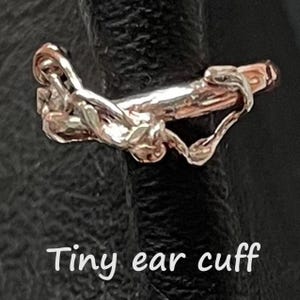May include: A small, silver-coloured ear cuff with an abstract design. The ear cuff is shown against a dark background. The text "Tiny ear cuff" is written in white cursive font at the bottom of the image.