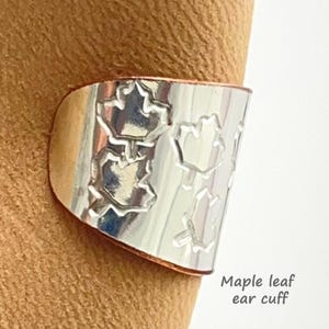 May include: A silver-toned metal ear cuff featuring a repeating embossed maple leaf pattern across its surface. The band has a smooth, reflective finish and includes the text Maple leaf ear cuff printed in the lower right corner of the image.
