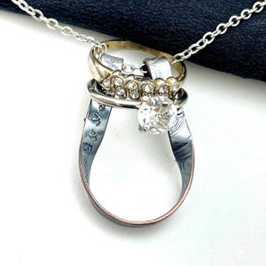 May include: A distinctive silver necklace composed of multiple rings. It features a prominent clear gemstone, a gold-coloured ring, and several silver rings. The necklace is suspended from a fine silver chain, displaying a mix of textures and designs.