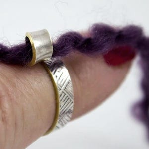 May include: A silver ring with a woven pattern and a gold band is shown. A silver and gold hair cuff is also visible, holding a strand of purple hair. The jewelry is displayed against a blurred background.