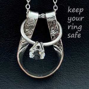 May include: A silver-toned metal ring holder pendant on a chain, featuring a textured, looped design that secures a ring with a clear, round-cut stone. The text "keep your ring safe" is printed in white on the right side of the image.