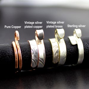 Może przedstawiać: Zbliżenie na cztery ręcznie robione pierścionki. Pierścionki wykonane są z czystej miedzi, posrebrzanej miedzi w stylu vintage, posrebrzanego mosiądzu w stylu vintage i srebra pr&oacute;by 925. Każdy pierścionek ma unikalny design i fakturę, z tekstem "Pure Copper", "Vintage silver plated copper", "Vintage silver plated brass" i "Sterling silver".