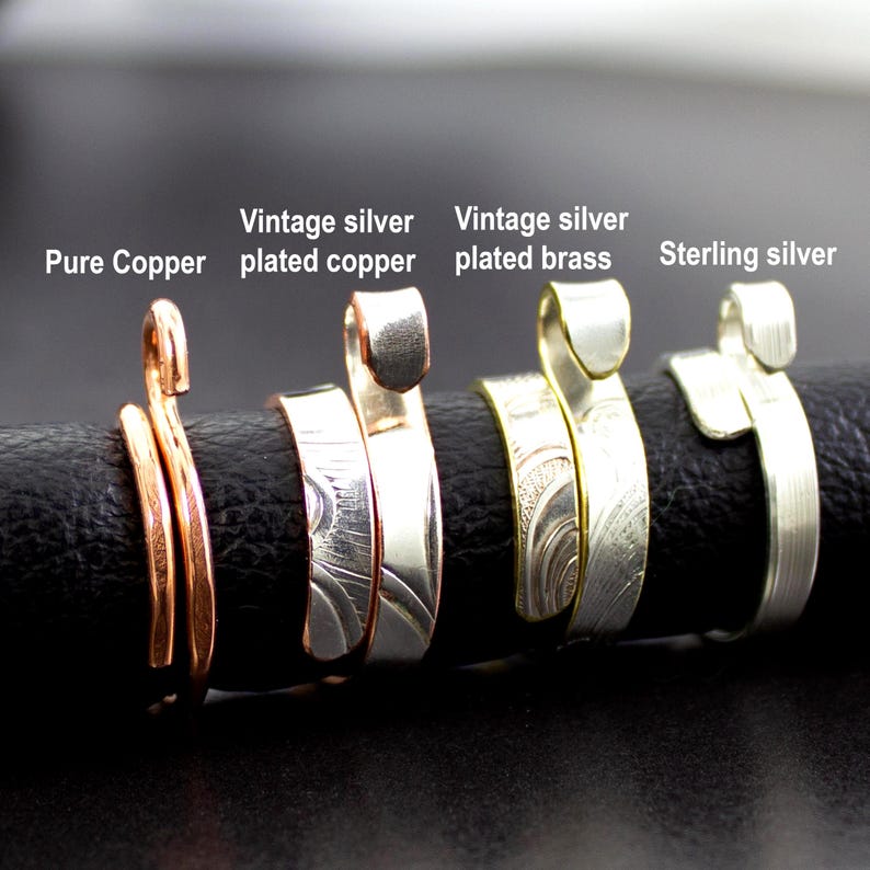 Może przedstawiać: Zbliżenie na cztery ręcznie robione pierścionki. Pierścionki wykonane są z czystej miedzi, posrebrzanej miedzi w stylu vintage, posrebrzanego mosiądzu w stylu vintage i srebra pr&oacute;by 925. Każdy pierścionek ma unikalny design i fakturę, z tekstem "Pure Copper", "Vintage silver plated copper", "Vintage silver plated brass" i "Sterling silver".