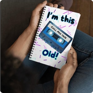 Puede incluir: Cuaderno de espiral con un diseño retro. La portada presenta un gráfico de cinta de cassette azul con las palabras "CHILL MIX TAPE" en la etiqueta. El texto "I'm this old!" está escrito en verde azulado y azul marino con acentos de rayos morados.