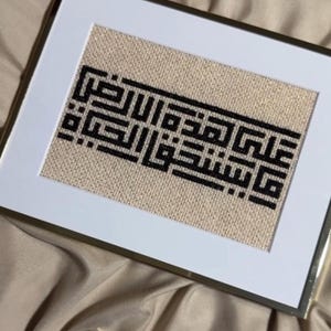 Op de afbeelding: Ingelijst kunstwerk met zwarte Arabische kalligrafie op een beige, getextureerde achtergrond. De kalligrafie is gecentreerd in een witte passe-partout en een goudkleurige lijst. Het kunstwerk wordt gepresenteerd op een gedrapeerde, lichtgeleurde stof.