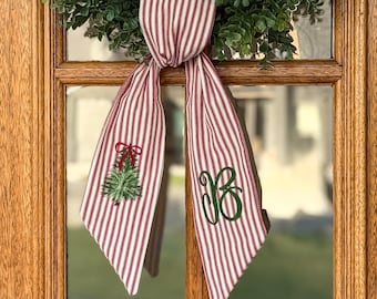 May include: A fabric ribbon featuring a red and white vertical striped pattern. One side displays an embroidered green Christmas tree with a red bow, while the other side features a large, dark green embroidered letter B in a script font.